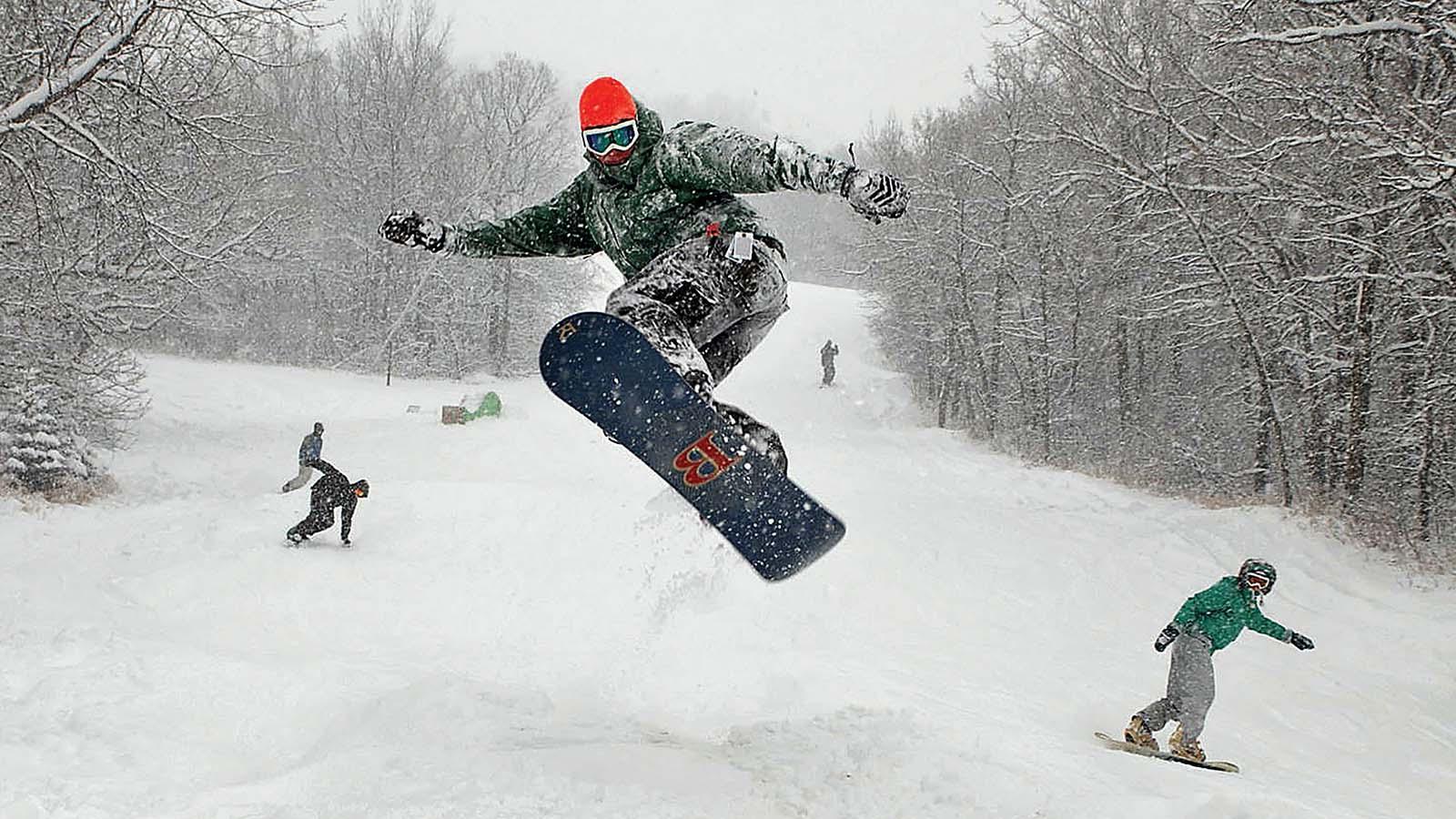 Snowboarder hitting a big jump at Huff Hills with several others snowboarding behind him