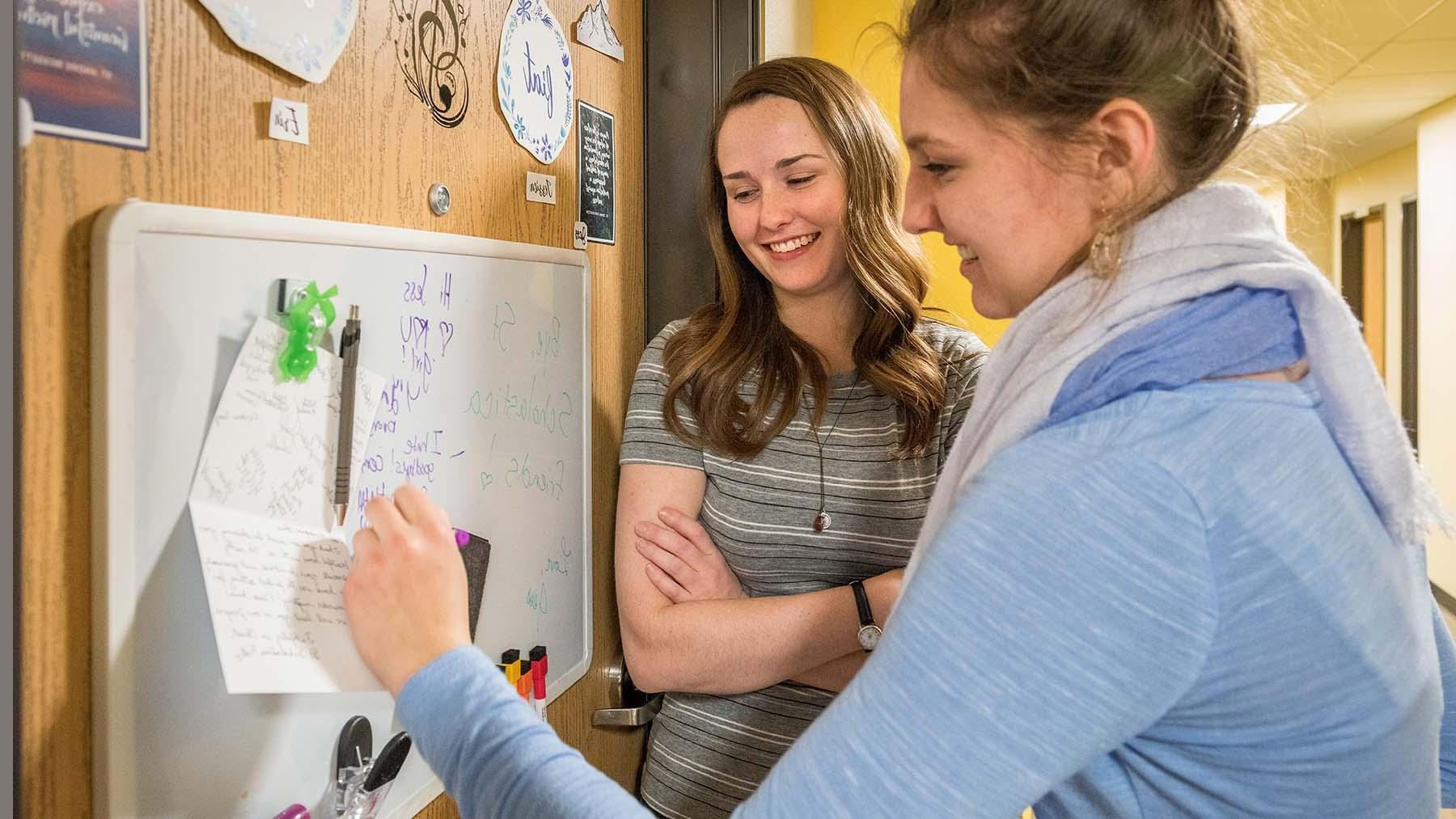 Two girls smiling and writing a message on a marker board on residence hall door